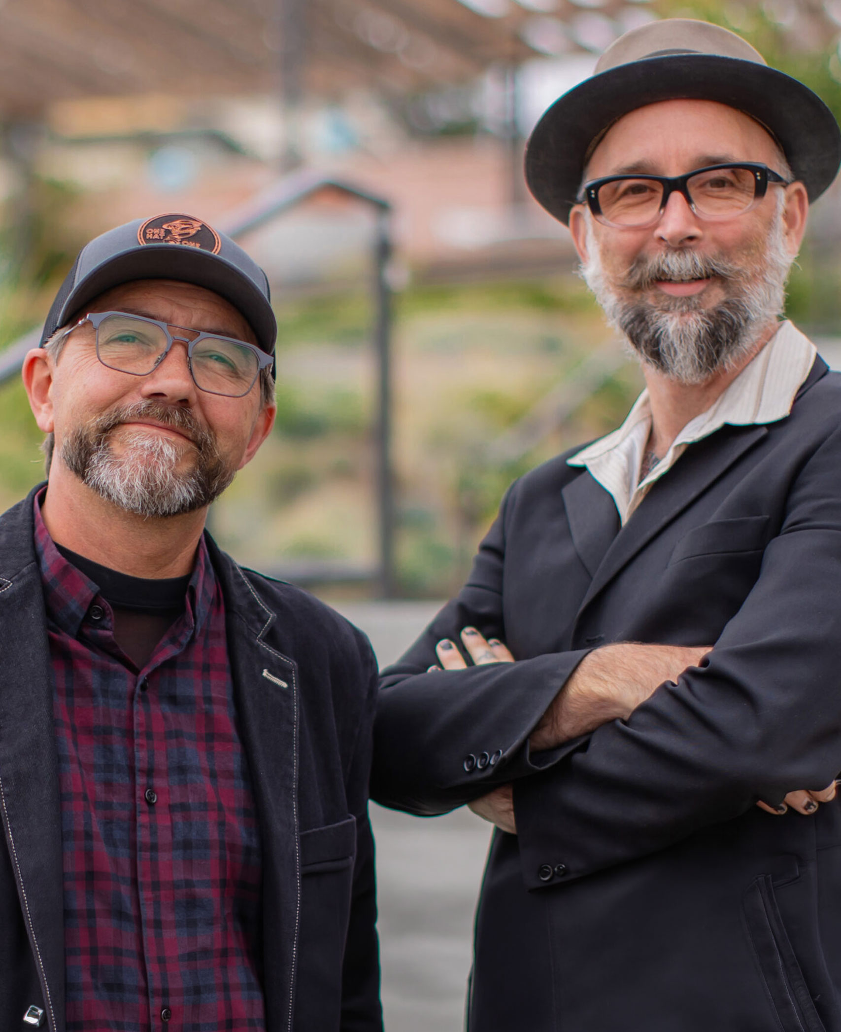 Two bearded men wearing glasses and hats posing together outdoors.