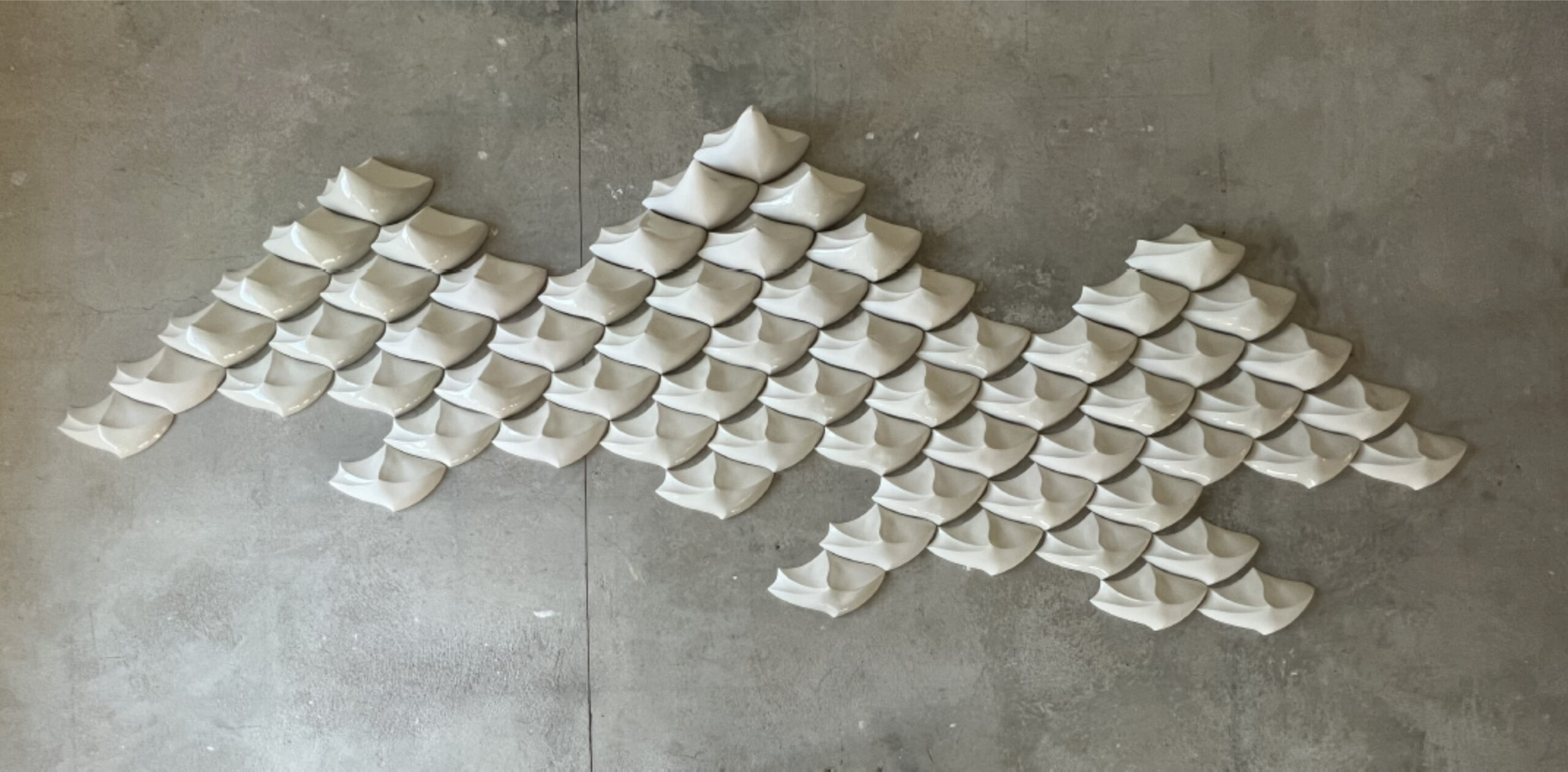 Wall installation of repeating white folded ceramic forms arranged in a flowing pattern